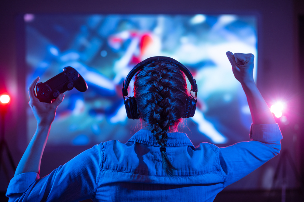 Girl in headphones and controller celebrating a win in front of a large gaming screen